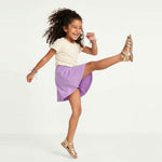 Child wearing a white top and purple skirt with gold sandals on a white background