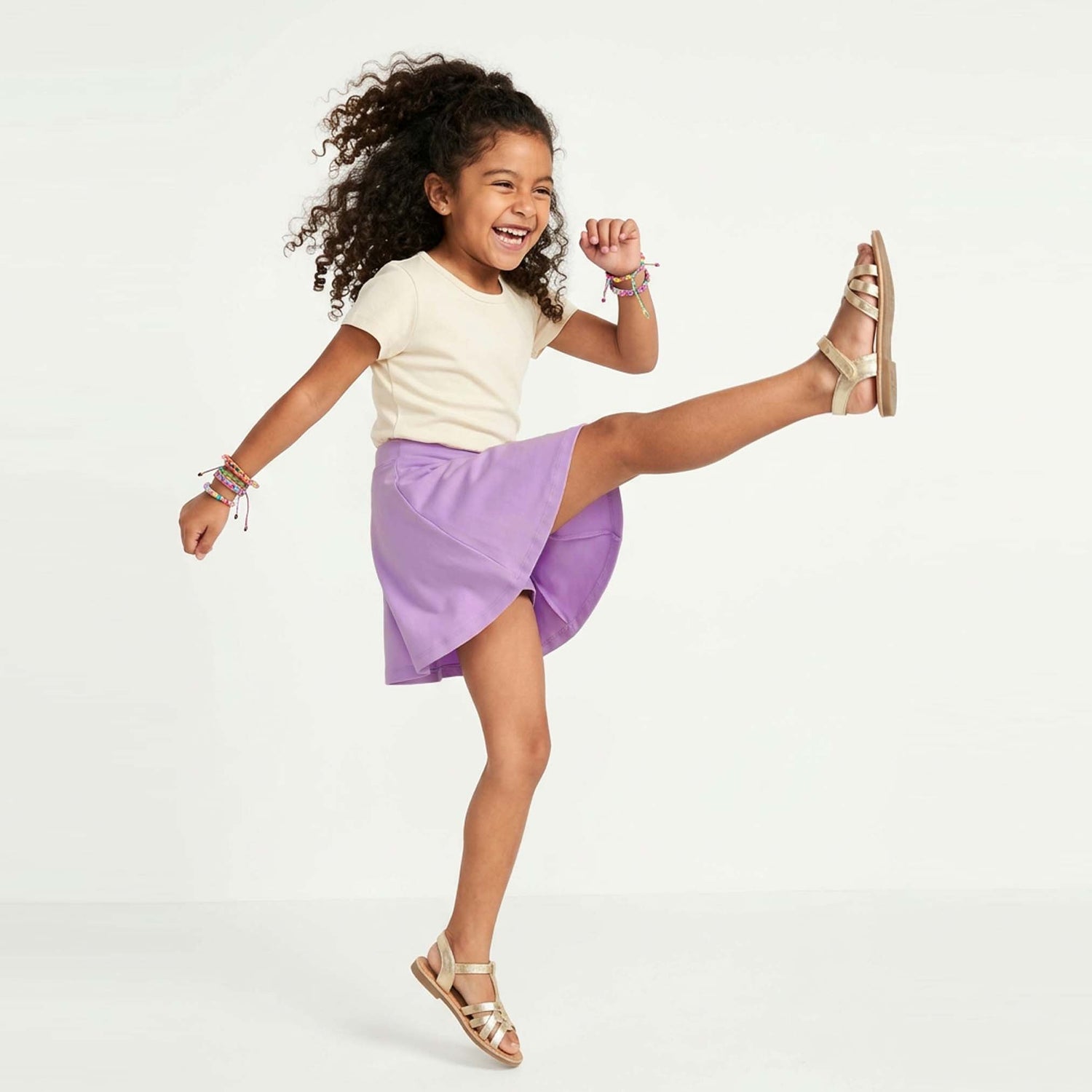 Child wearing a white top and purple skirt with gold sandals on a white background