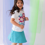 young girls wearing a white tee shirt with a floral print and a turquoise skirt against a pastel background