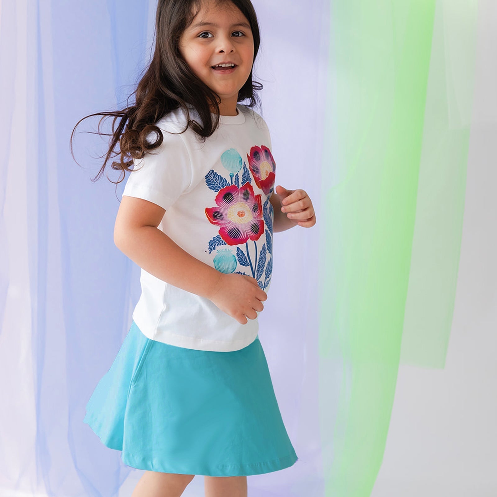 young girls wearing a white tee shirt with a floral print and a turquoise skirt against a pastel background