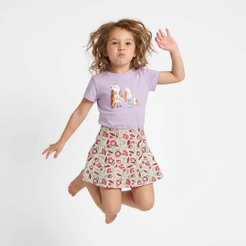 Young girl jumping. She is wearing a Mightly 100% organic cotton graphic tee with a colorful bird print and a floral skirt. 