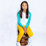 a girl sitting on a wooden stool wearing a teal and white long sleeve baseball tee and yellow pants
