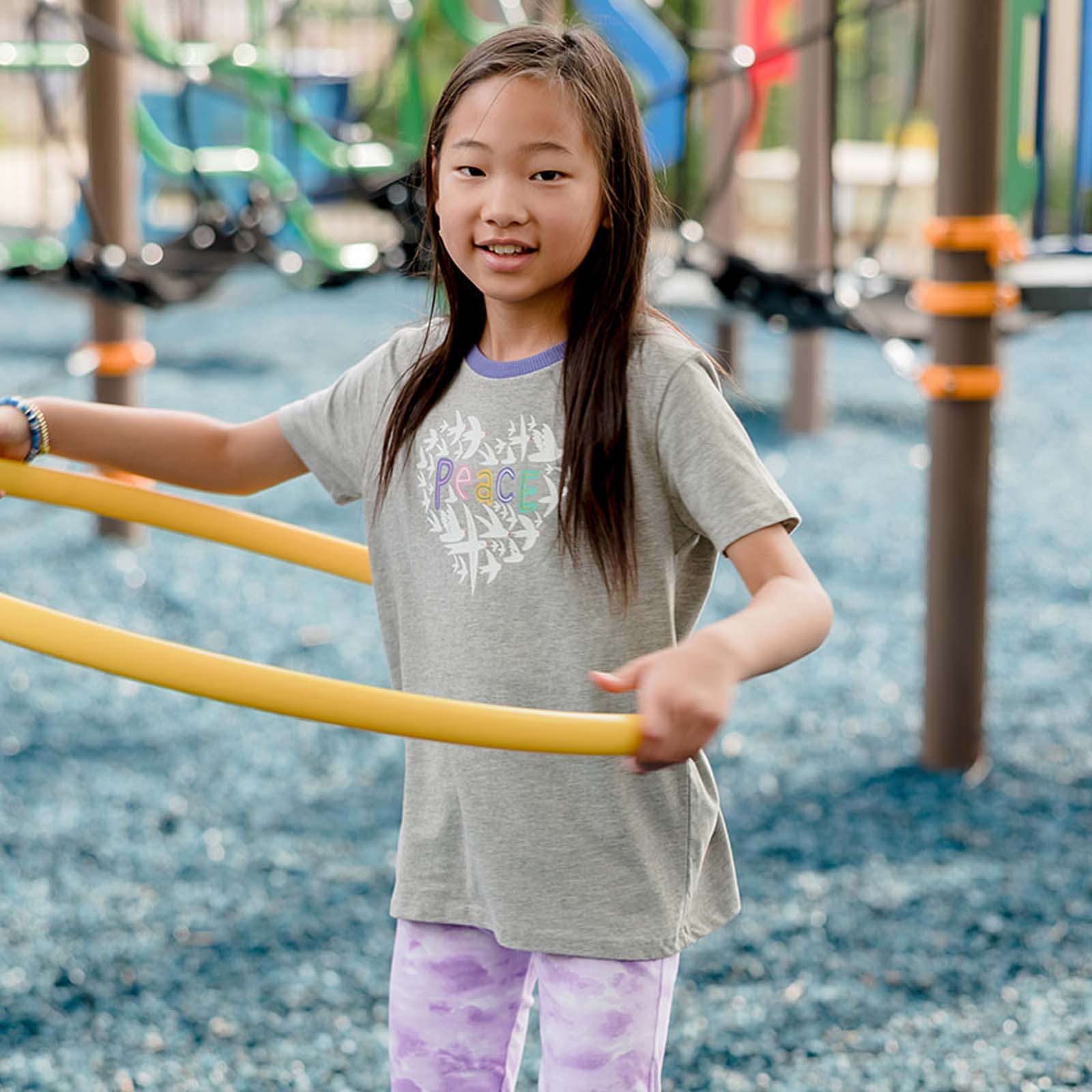 Young girl playing with a yellow hula hoop on a playground. Wearing a gray tee shirt with peace printed on it.
