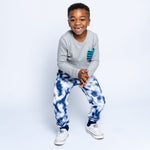 A young boy wearing a grey long sleeve tee with a striped pocket and tie dye sweatpants